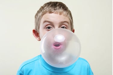 Child with blue shirt blowing a large pink bubble from bubble gum. 