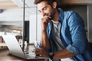 Man in white shirt and blue over button up shirt looking at a laptop. 