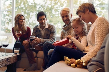 Family sitting around visiting. Including four adults and one child. Child is opening a present while some adults are drinking wine on a clean couch. 