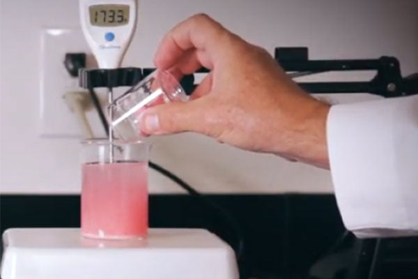 Hand pouring liquid into a beaker full of pink liquid. 
