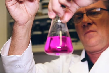 Person in white lab coat looking at pink chemical in beaker. 